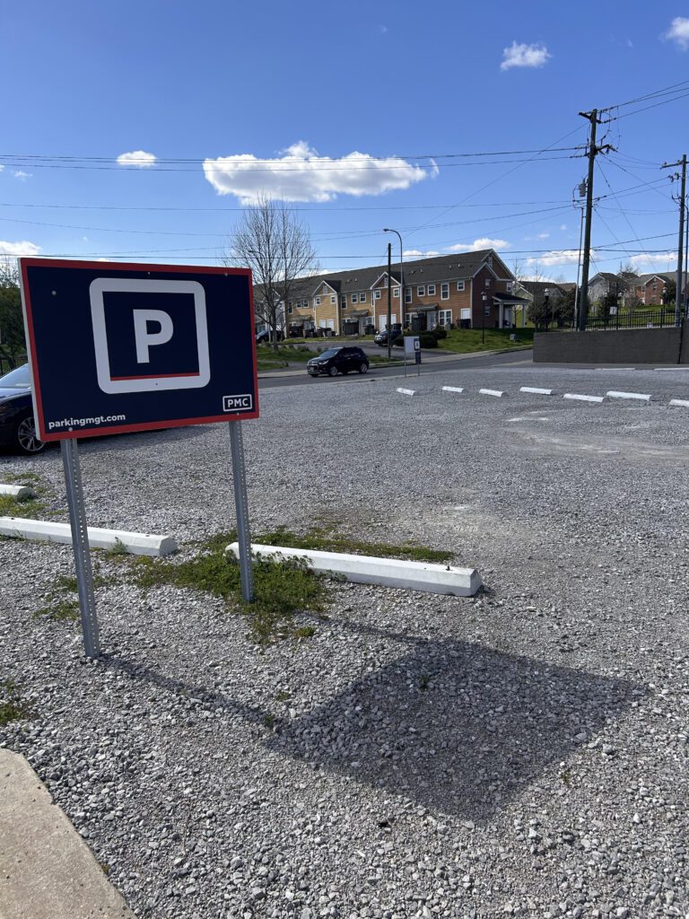A mostly empty gravel parking lot with a large PMC parking sign in the foreground. There are a few cars and houses in the background under a blue sky with scattered clouds.