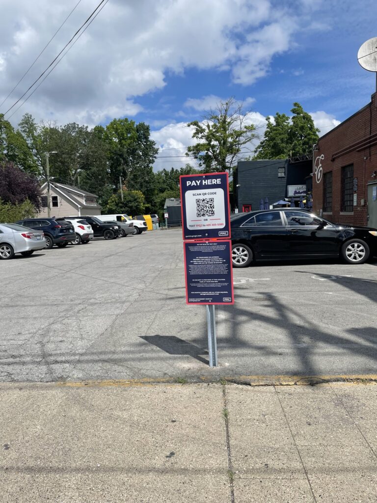 A parking lot with several parked cars and a “Pay Here” sign featuring QR code payment instructions stands on the sidewalk under a partly cloudy sky. Trees and buildings are visible in the background.
