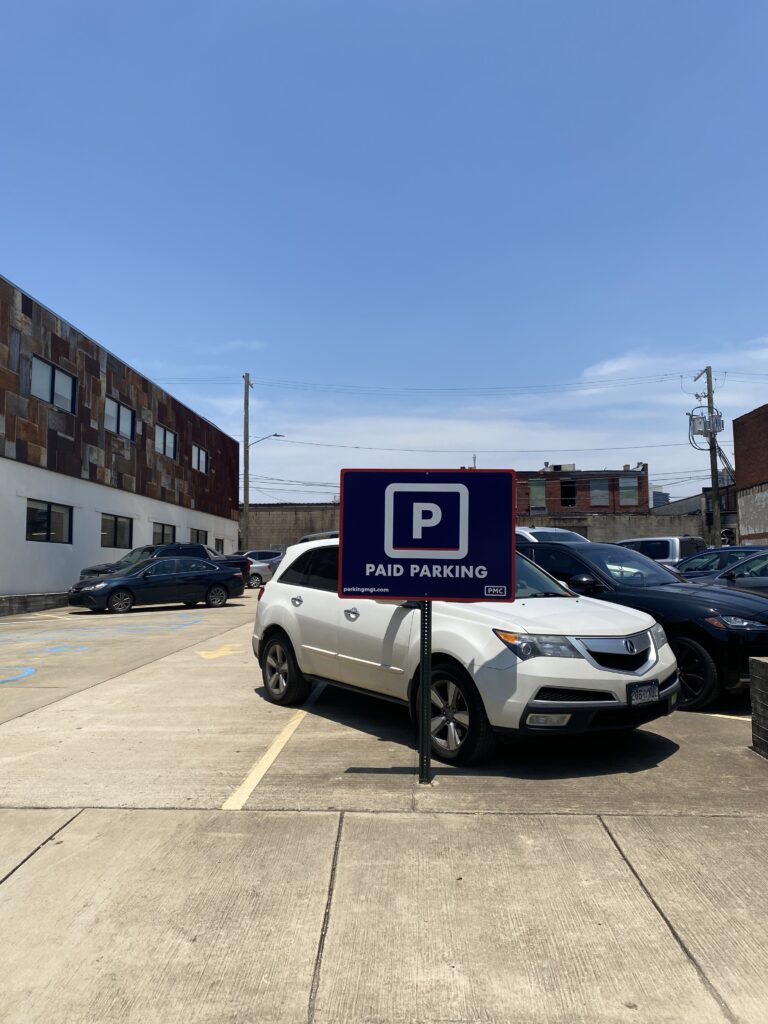 A white SUV is parked in a lot next to a sign that reads "Paid Parking." Other cars and a building are visible in the background under a clear blue sky.