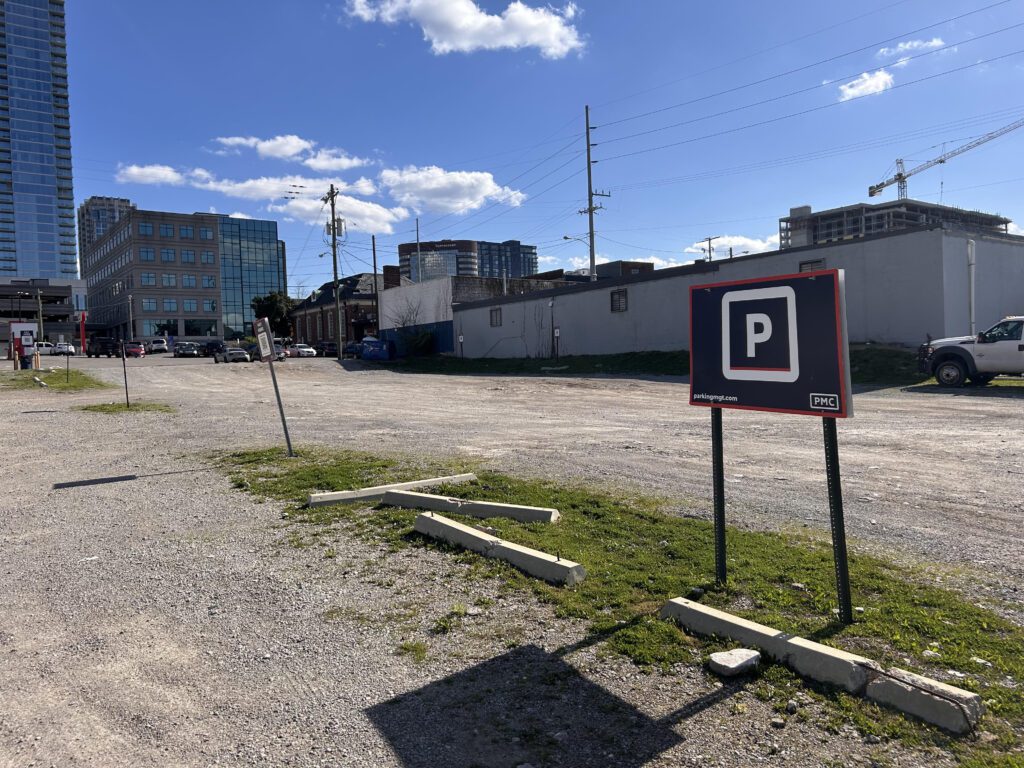 A gravel parking lot with a black and white "P" parking sign in the foreground, several parked cars, and buildings in the background under a blue sky with scattered clouds.