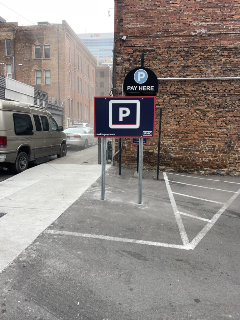 Parking lot with "Pay Here" and "P" parking signs near a brick building; a sidewalk and parked van are on the left, with empty parking spaces and faded lines visible on the pavement.