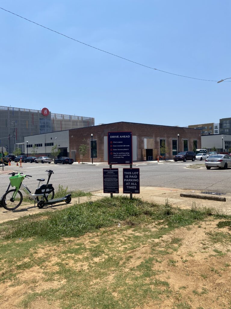 A parking lot with a sign displaying parking rates and rules stands in the foreground. A bike and scooter are parked on the left, with a brick building and other modern buildings in the background under a clear blue sky.