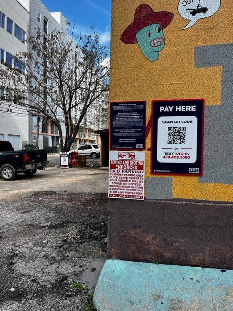 A colorful mural with a talking cartoon face is on a wall near multiple parking signs, including a QR code payment sign and a towing warning; parked vehicles and buildings are visible in the background.