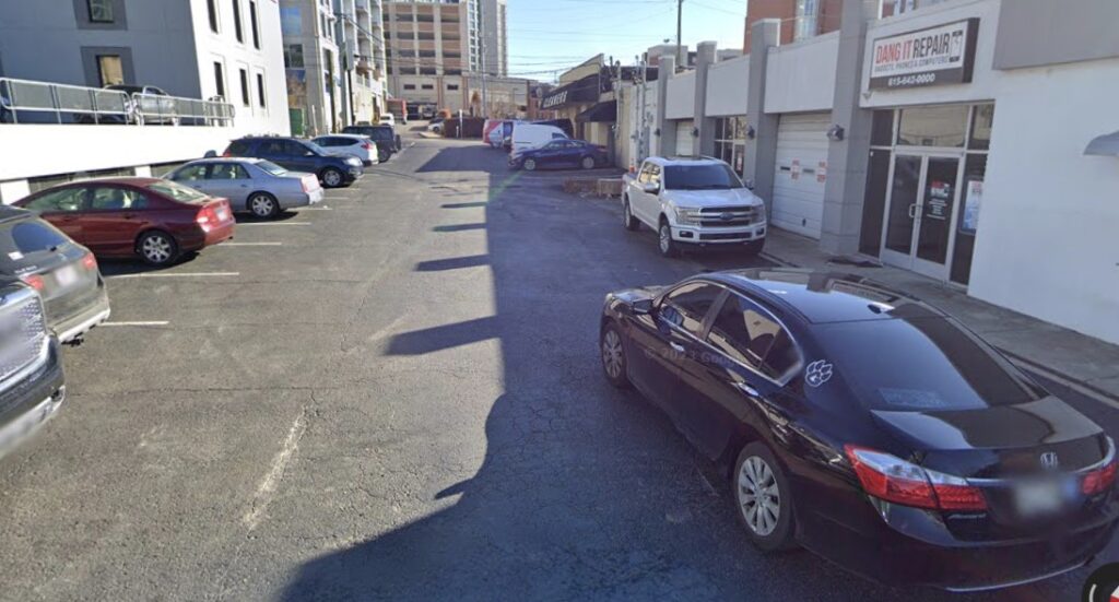 A parking lot beside a row of buildings, with several parked cars including a black sedan in the foreground and a white truck near an auto repair shop named "Davis Repair." Urban buildings and a van are visible in the background.