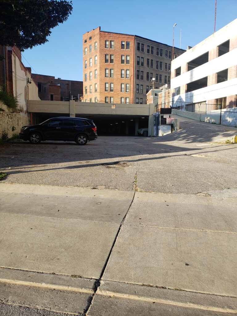 A parked black SUV sits near a parking garage entrance at the base of a multi-story brick building, with a white multi-level parking structure on the right and clear blue sky above.