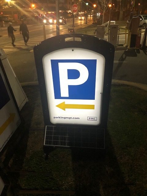 Illuminated parking sign with a large white "P" on a blue background and a yellow arrow pointing right. The sign sits on grass near a street at night with traffic and people in the background.