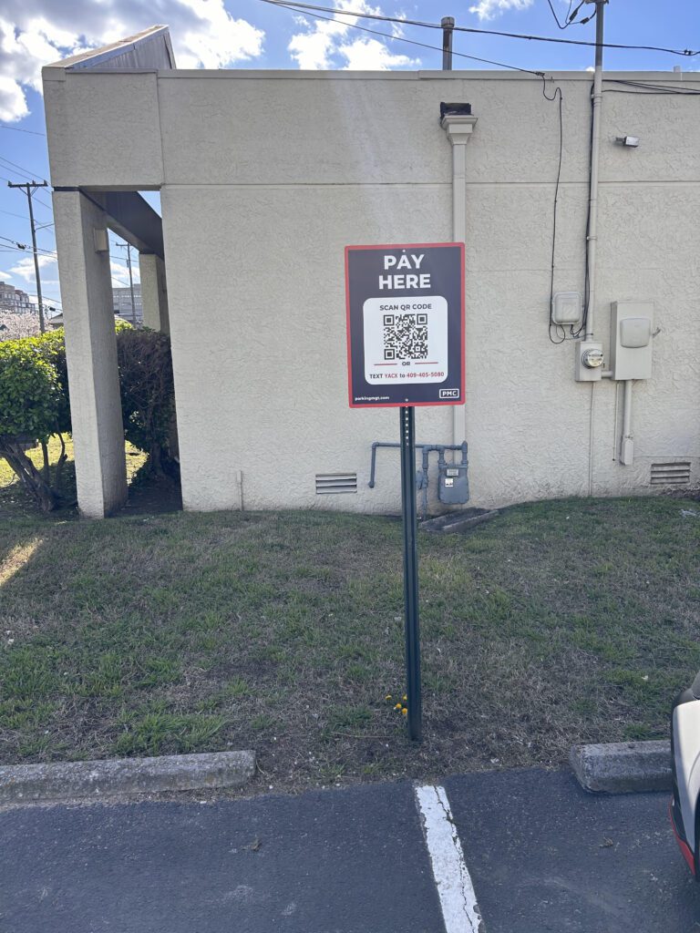 A sign in a parking lot reads "PAY HERE" with a QR code for payment instructions. The sign is on a post beside a building, on a grassy patch near a parking space under a partly cloudy sky.