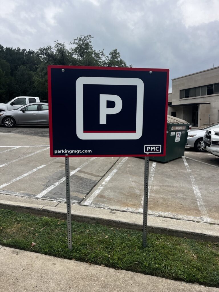 A large sign with a white "P" on a dark blue background indicates parking. The sign stands on grass near a parking lot with cars, a dumpster, and buildings in the background.