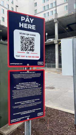 A parking payment sign instructs users to scan a QR code or text a number to pay. The sign is posted on a pole in an outdoor parking lot near a building and other infrastructure.
