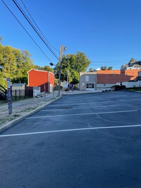 A mostly empty parking lot with marked spaces, surrounded by buildings, including a red house and a brick building, on a clear, sunny day. Trees and power lines are visible in the background.