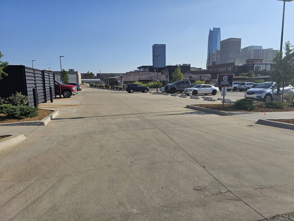 A mostly empty parking lot on a sunny day with a few cars parked; tall city buildings are visible in the background under a clear blue sky.