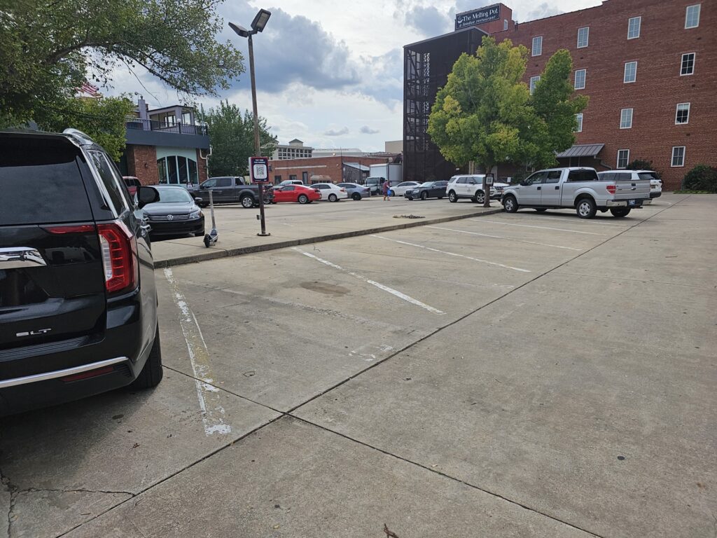 A nearly empty parking lot with a few cars parked along the edges, bordered by brick buildings and trees under a cloudy sky. A black SUV is visible in the foreground on the left.