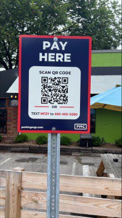 A sign with "Pay Here" at the top features a QR code for scanning, and instructions to text "MF2Y" to 469-405-5080 for payment. The sign is outdoors near a parking lot and wooden fence.