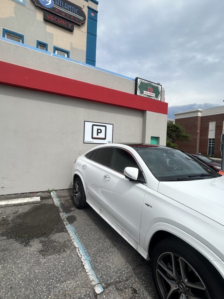 A white car is parked at an angle in front of a gray wall with a PMC parking sign.