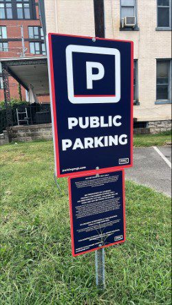 A sign in grassy area reads "Public Parking" in bold white letters on a dark background, indicating a parking area. There are buildings and part of a parking lot visible in the background.