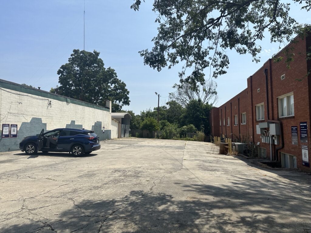 A nearly empty parking lot with one parked blue SUV on the left. The lot is bordered by a white building on the left and a red brick building on the right, with trees and greenery in the background.