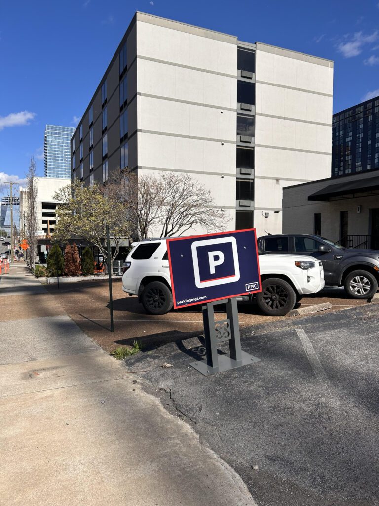 A parking lot with several parked cars next to a modern, multi-story building. A large sign with a white "P" on a blue background indicates parking. Trees and a sidewalk are also visible under a clear blue sky.