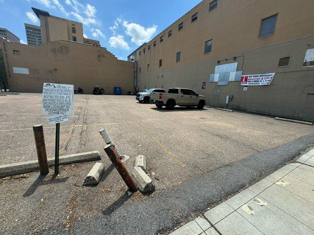 Nearly empty urban parking lot with a few parked vehicles along the back wall. Several damaged concrete barriers and leaning poles are in the foreground. Large tan buildings surround the lot under a partly cloudy sky.