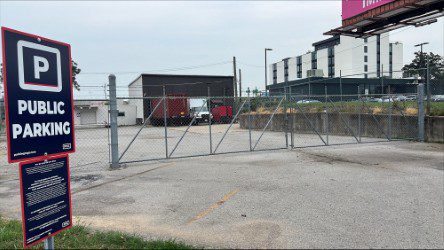 A fenced and gated parking lot with a "Public Parking" sign in the foreground. In the background, there are buildings, parked vehicles, and a cloudy sky.