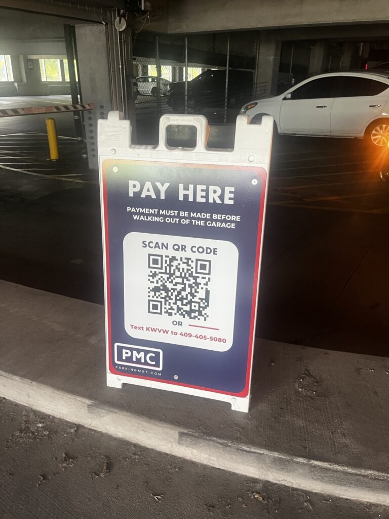 A sign in a parking garage reads "Pay Here. Payment must be made before walking out of the garage." It has a QR code for payment and a phone number, with the PMC logo at the bottom. A white car is parked in the background.