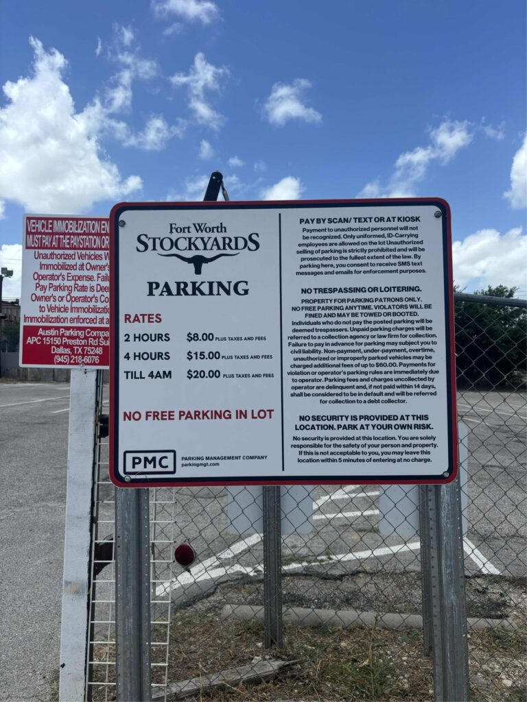 A sign at Fort Worth Stockyards parking lot lists rates: 2 hours $8, 4 hours $15, until 4am $20. It states no free parking, no trespassing, no security, and gives payment and parking rules. Fence and sky in background.