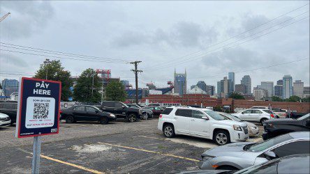 A parking lot with several cars and a "Pay Here" sign in the foreground; the Nashville city skyline, including the AT&T Building, is visible in the background under a cloudy sky.