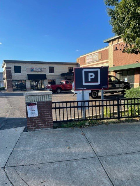A parking lot with a black parking sign in the foreground, a sidewalk, a metal fence, two parked trucks, and a two-story brick and tan building under a clear blue sky.