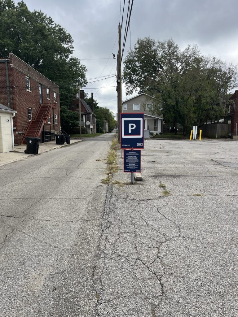 A parking lot with a blue sign marked “P” for parking stands in the center. The lot is mostly empty, bordered by houses and trees under a cloudy sky. Grass grows through cracks in the pavement.
