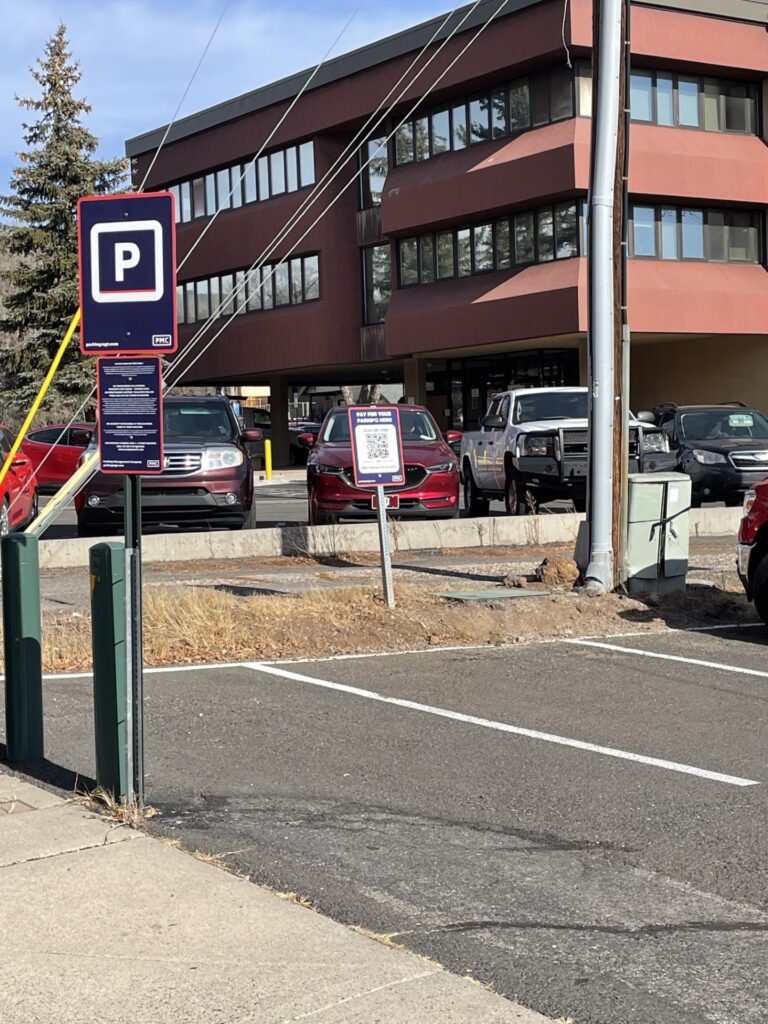 A parking lot with several parked cars near a brown office building. A parking sign and a payment information sign with a QR code are visible in the foreground. Some grass and a sidewalk border the lot.