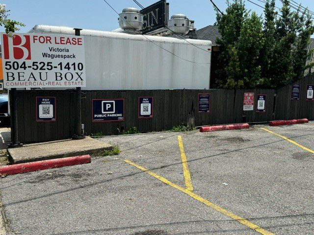 A parking lot with faded yellow lines, red curbs, and a wooden fence displaying several "Pay Here" and "Public Parking" signs. A "For Lease" sign with a phone number hangs on the fence. Trees and a building are in the background.