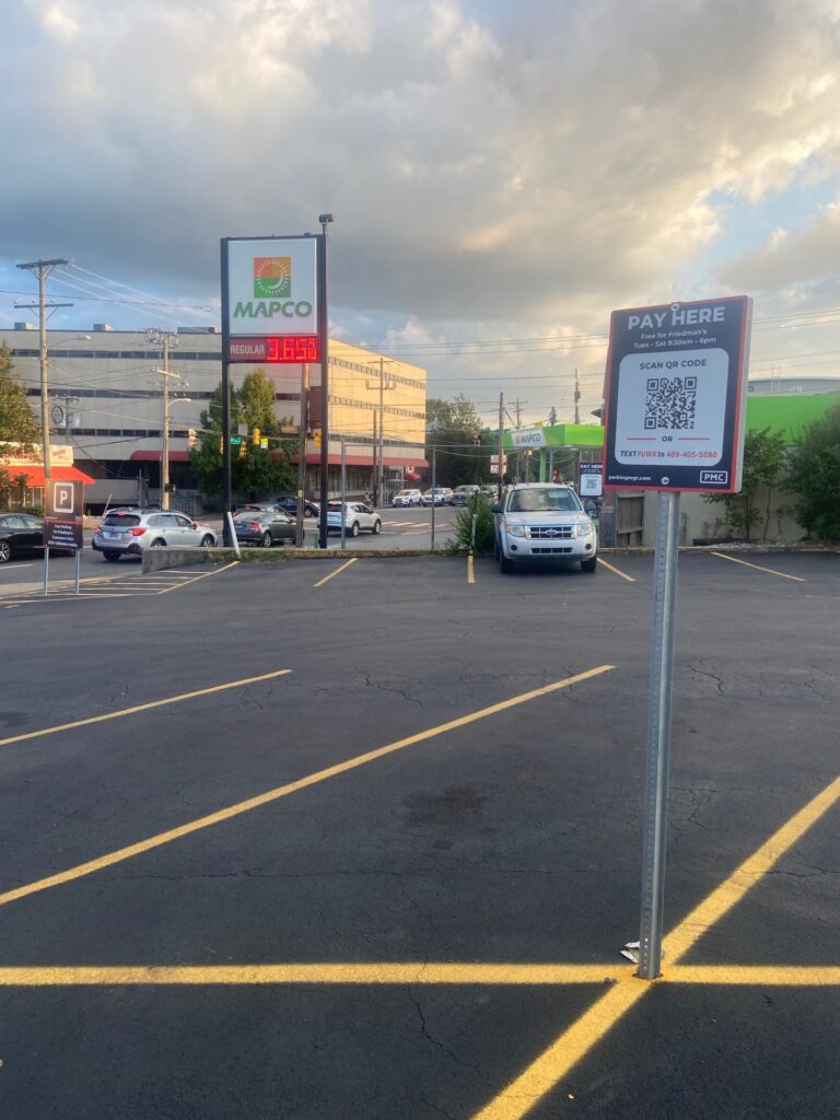 A mostly empty parking lot with yellow lines, a pay station sign featuring a QR code, and a MAPCO gas station with fuel prices in the background under a cloudy sky.