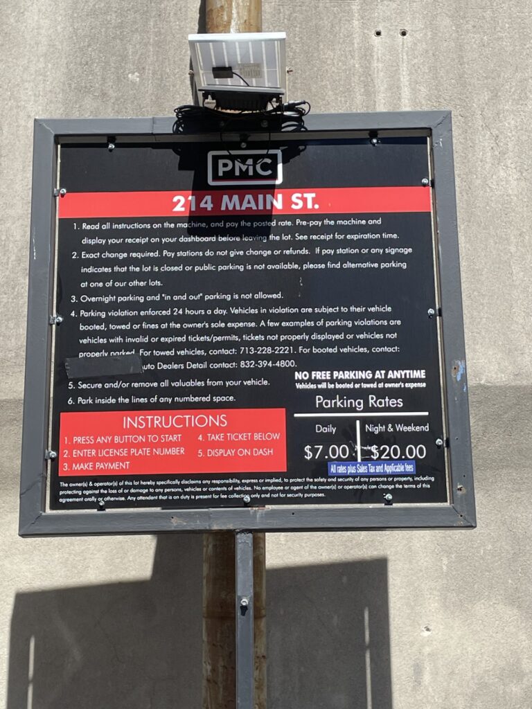A black and red PMC parking sign for 214 Main St. lists parking rules, instructions, parking rates ($7 daily, $20 night/weekend), and a warning that unauthorized or unpaid vehicles will be towed at the owner’s expense.