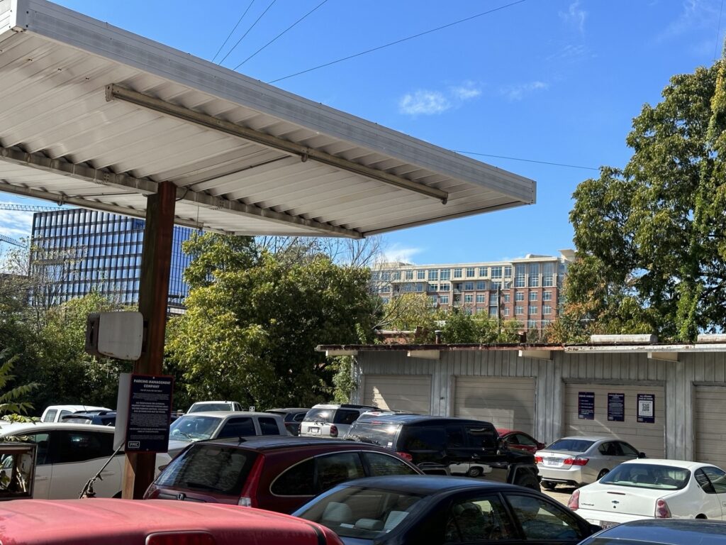 A parking lot filled with cars next to a white-roofed structure, with trees and a multi-story building in the background under a blue sky.