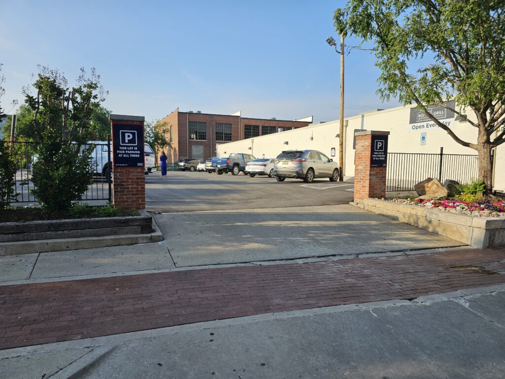 Entrance to a parking lot with several parked cars, flanked by brick pillars and “Public Parking” signs. A few trees and flower beds line the edges, and a person walks in the distance.
