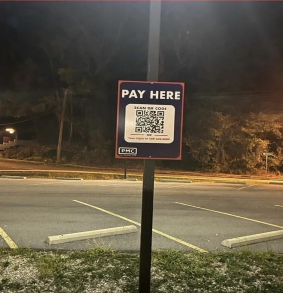 A sign in an empty parking lot at night reads "PAY HERE" with a QR code below it for payment. The area is dimly lit, and trees are visible in the background.