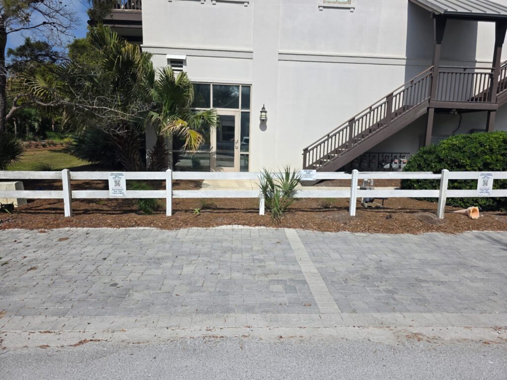A white fence lines a sidewalk in front of a light-colored building with stairs on the right. Sparse plants and small trees grow in the mulch beside the fence. The paved area in the foreground appears to be a parking spot.