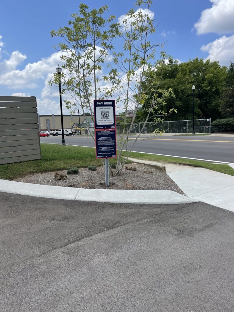 A “Pay Here” parking sign with a QR code stands next to a small tree and landscaped area by a road on a sunny day, with a building, cars, fence, and blue sky with clouds in the background.
