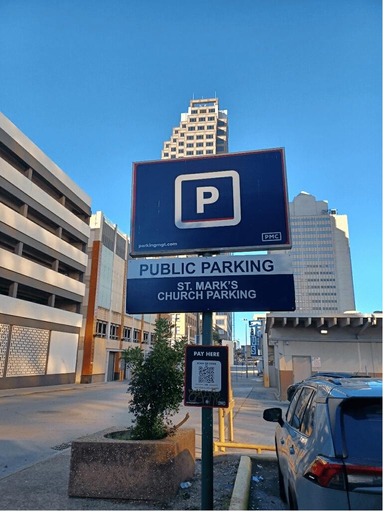 A public parking sign for St. Mark's Church Parking stands on a city street, with a tall building and parked cars visible in the background under a clear blue sky.