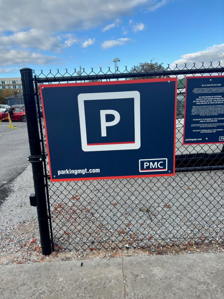 A sign on a chain-link fence displays a large white letter "P" in a square, indicating parking. Below, "parkingmgt.com" and "PMC" appear on a navy background. Cars and cloudy sky are visible in the background.