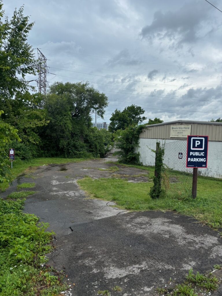A cracked and uneven asphalt driveway leads past trees and a metal building with a “Public Parking” sign, under a cloudy sky. The area appears overgrown with grass and weeds along the edges.