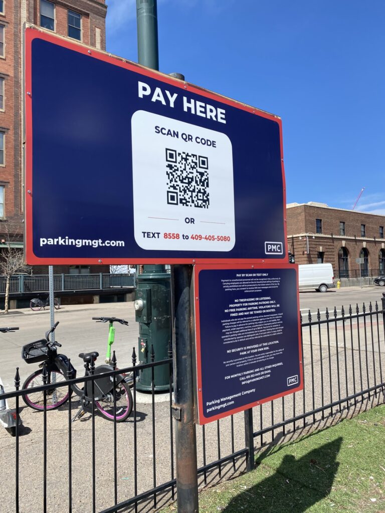 A parking payment sign outdoors reads "Pay Here" and displays a QR code for payment, a phone number, and instructions. Two bikes are parked nearby; buildings and a fence are visible in the background under a blue sky.