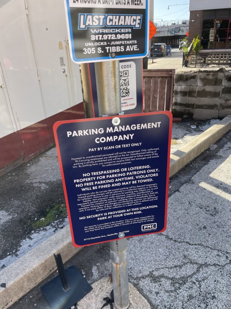 A sign on a pole in a parking lot displays parking rules, payment instructions (by scan or text), warnings about trespassing, and states parking is at your own risk. Nearby, a "Last Chance Wrecker" sign is visible above.