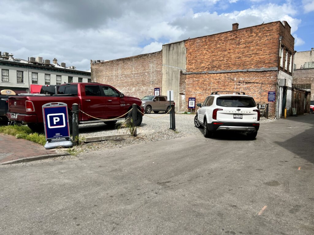 A small gravel parking lot with a red pickup truck and a white SUV parked. There are several PMC parking signs with instructions for digital payment. There is a brick building with faded paint in the background under a partly cloudy sky.