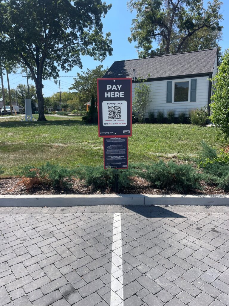 A "Pay Here" sign with a QR code stands at the edge of a paved parking lot, with a grassy area, shrubs, and a white house in the background.