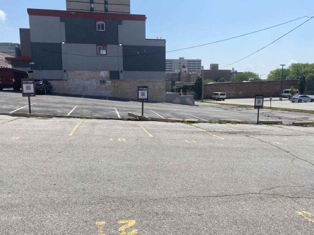 A mostly empty parking lot with numbered spaces and signs, situated near a gray building with red trim under a clear blue sky. A few vehicles are parked along the lot’s edge.