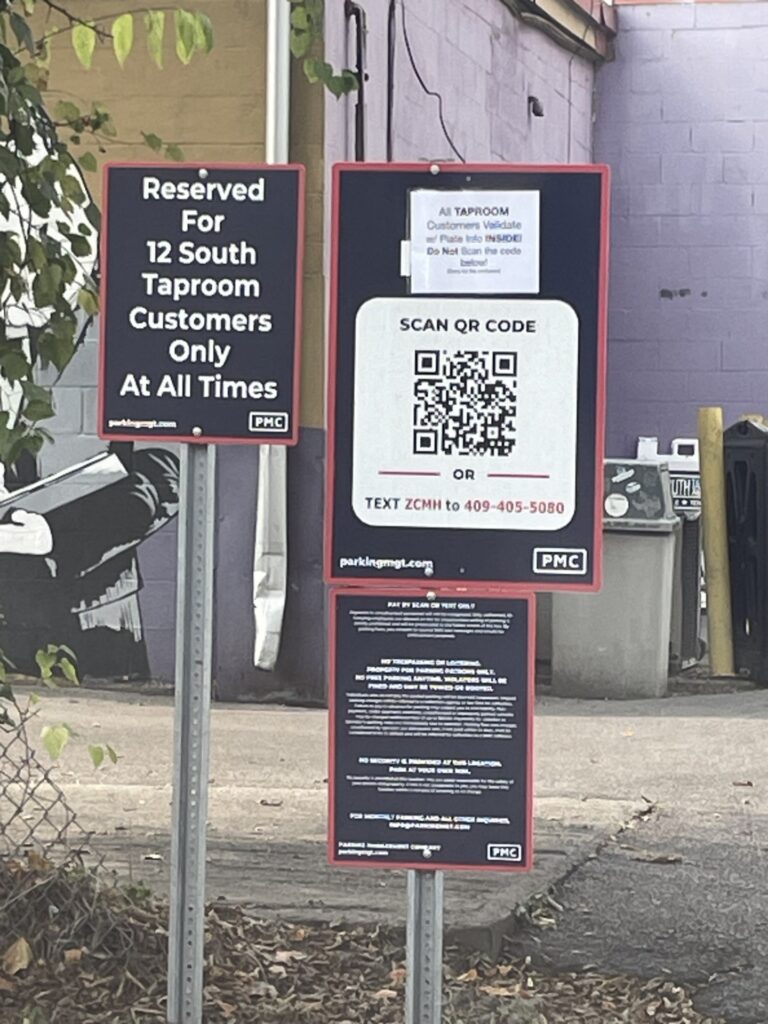 Signs in a parking lot indicate that parking is reserved for 12 South Taproom customers only. One sign has a QR code and text instructions for payment or access. A mural and trash bins are visible in the background.