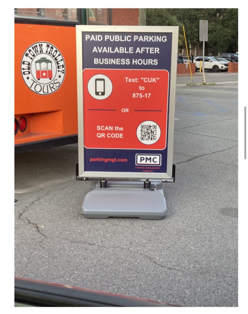 A sign in a parking lot reads: “Paid public parking available after business hours. Text ‘CUK’ to 875-17 or scan the QR code.” There’s a QR code and a nearby orange trolley with “Old Town Trolley Tours” written on it.