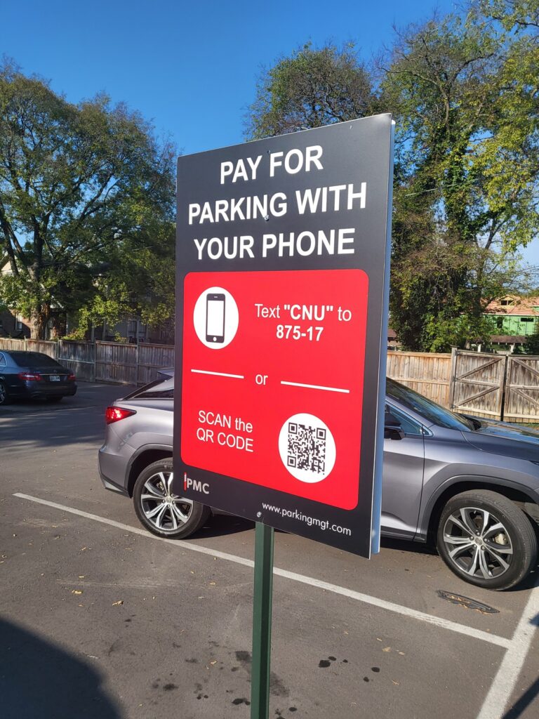 A parking lot sign instructs people to pay for parking with their phones by texting "CNU" to 875-17 or scanning a QR code. Several cars and trees are visible in the background.