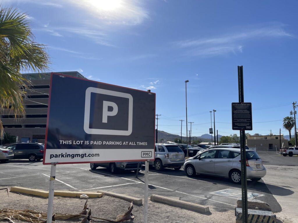 A large PMC sign in a parking lot reads “This lot is paid parking at all times." Several vehicles are parked in the background, with a clear blue sky and a few palm trees visible.