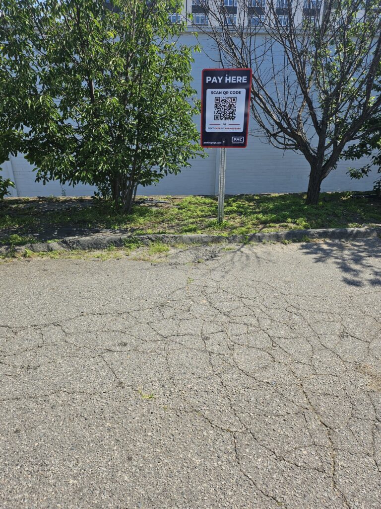 A “Pay Here” sign for parking stands on a cracked pavement near some trees and a white wall. The sign includes a QR code for payment and instructions. The area is sunny with patches of grass nearby.